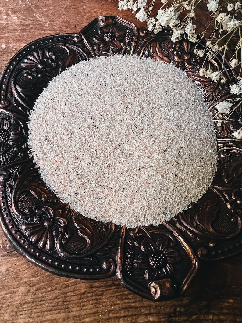 May include: A white, fine-grained powder sits in a decorative, ornate, bronze-colored dish with floral designs. The dish is on a wooden surface.