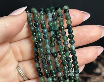 May include: A strand of green moss agate beads, with a mottled green and black pattern, strung on a clear elastic cord. The beads are round and smooth, and the strand is approximately 6 inches long.