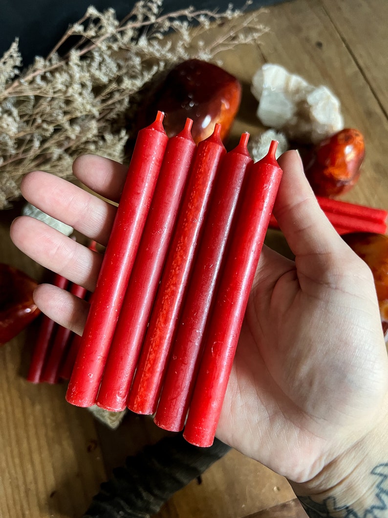 May include: A hand holding six red taper candles. The candles are cylindrical with a slightly textured surface and a small wick at the top. The background includes dried plants and various stones.