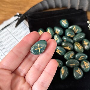 May include: A set of 25 green agate rune stones with gold lettering. The stones are in a velvet pouch and are being held in a hand. The runes are used for divination and meditation.