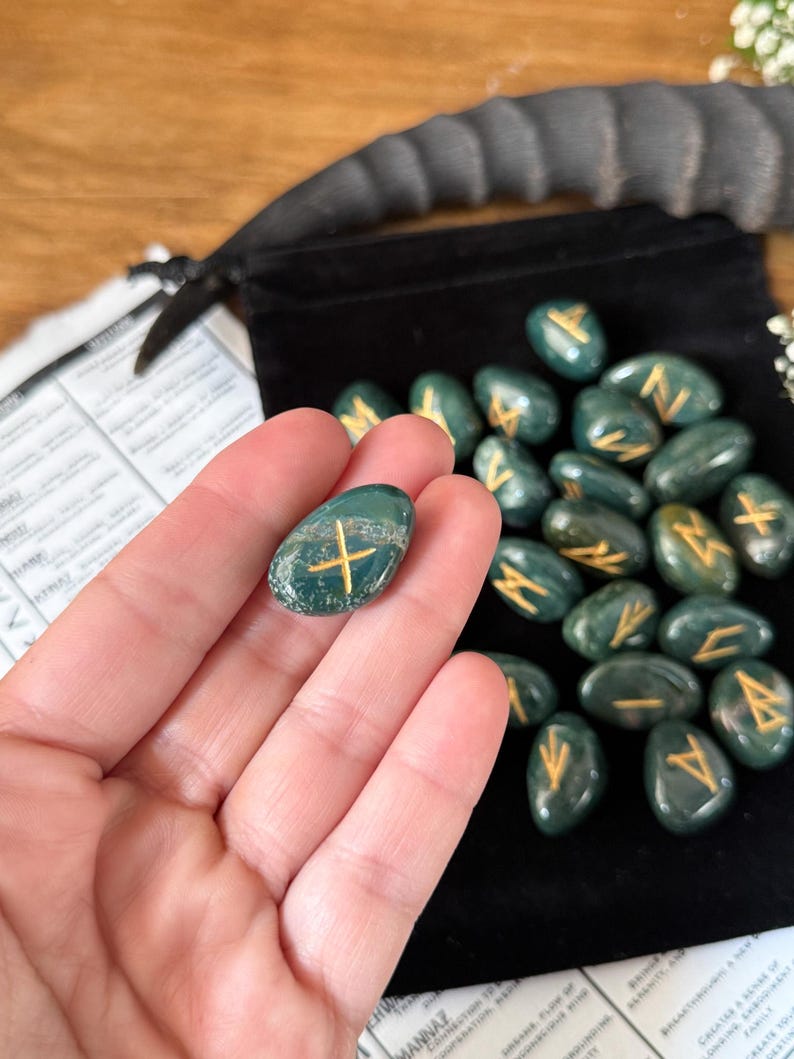 May include: A set of 25 green agate rune stones with gold lettering. The stones are in a velvet pouch and are being held in a hand. The runes are used for divination and meditation.
