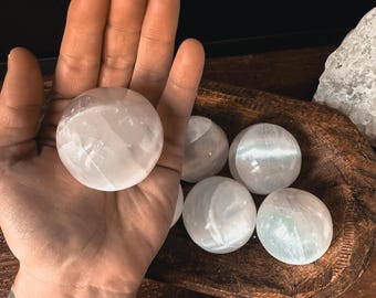 Op de afbeelding: Een verzameling van zes witte seleniet bollen, waarvan sommige in een houten kom zitten. De bollen zijn glad en hebben een doorschijnende uitstraling.