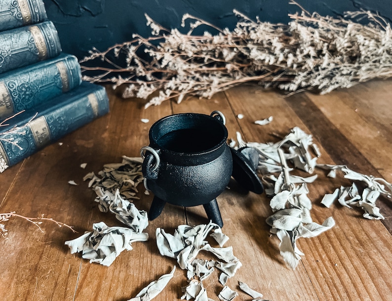 May include: A small black cast iron cauldron with a lid and three legs. The cauldron is sitting on a wooden surface with white sage leaves scattered around it.