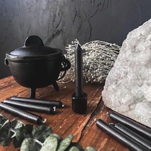 May include: A black cast iron cauldron with a lid, surrounded by black taper candles, a crystal, and dried flowers. The items are arranged on a rustic wooden surface, creating a mystical aesthetic.