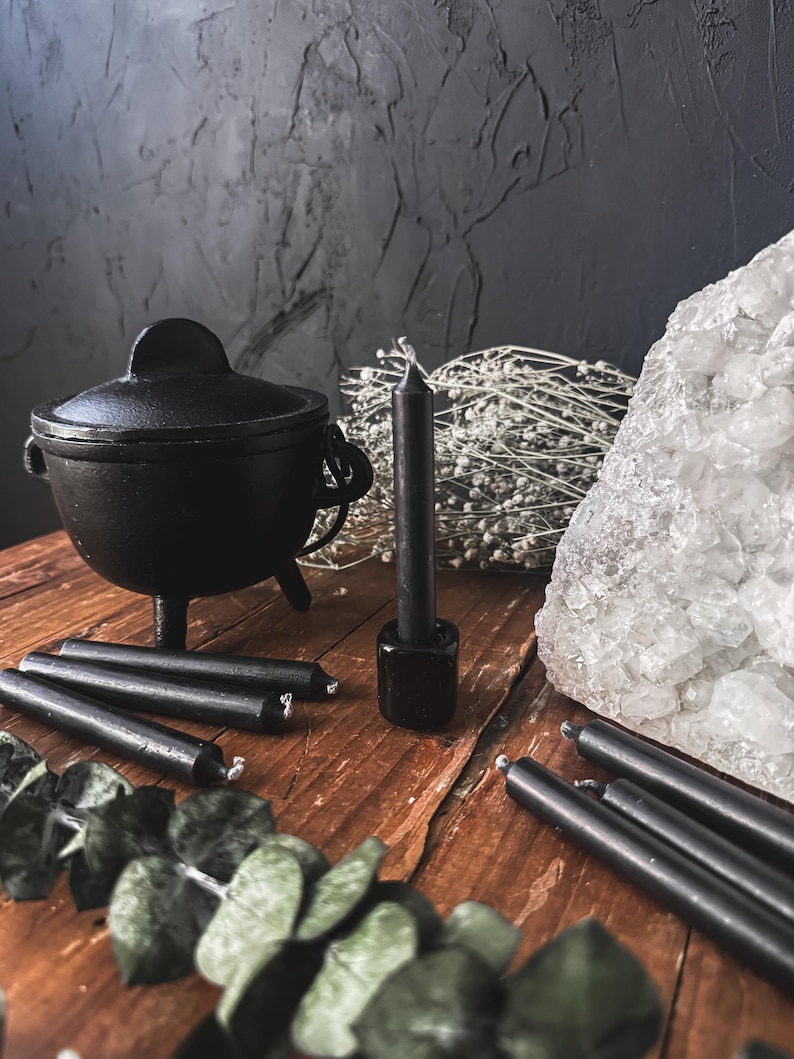 May include: A black cast iron cauldron with a lid, surrounded by black taper candles, a crystal, and dried flowers. The items are arranged on a rustic wooden surface, creating a mystical aesthetic.
