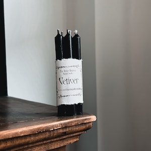 May include: A bundle of four black ritual candles with white wicks, wrapped with a label that reads "Vetiver" and "The Stone Modern Ritual Candles". The candles are displayed on a dark wooden surface.