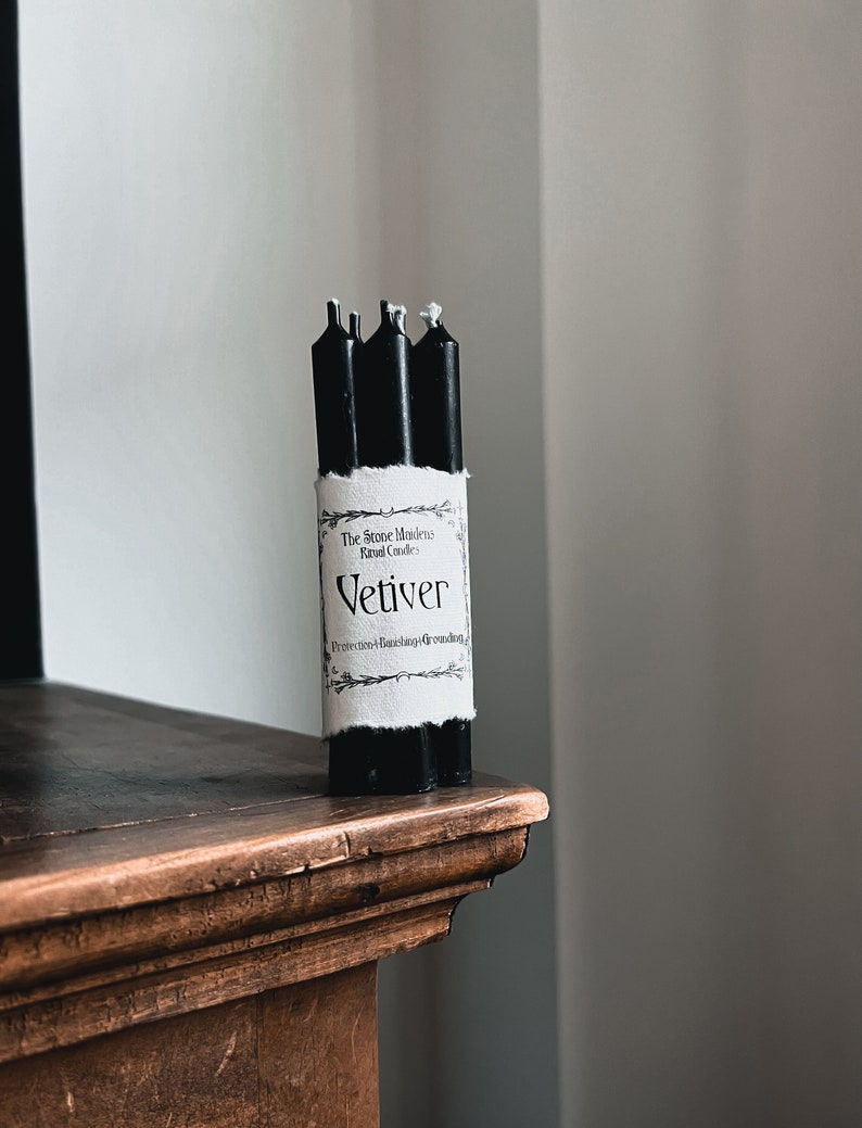May include: A bundle of four black ritual candles with white wicks, wrapped with a label that reads "Vetiver" and "The Stone Modern Ritual Candles". The candles are displayed on a dark wooden surface.
