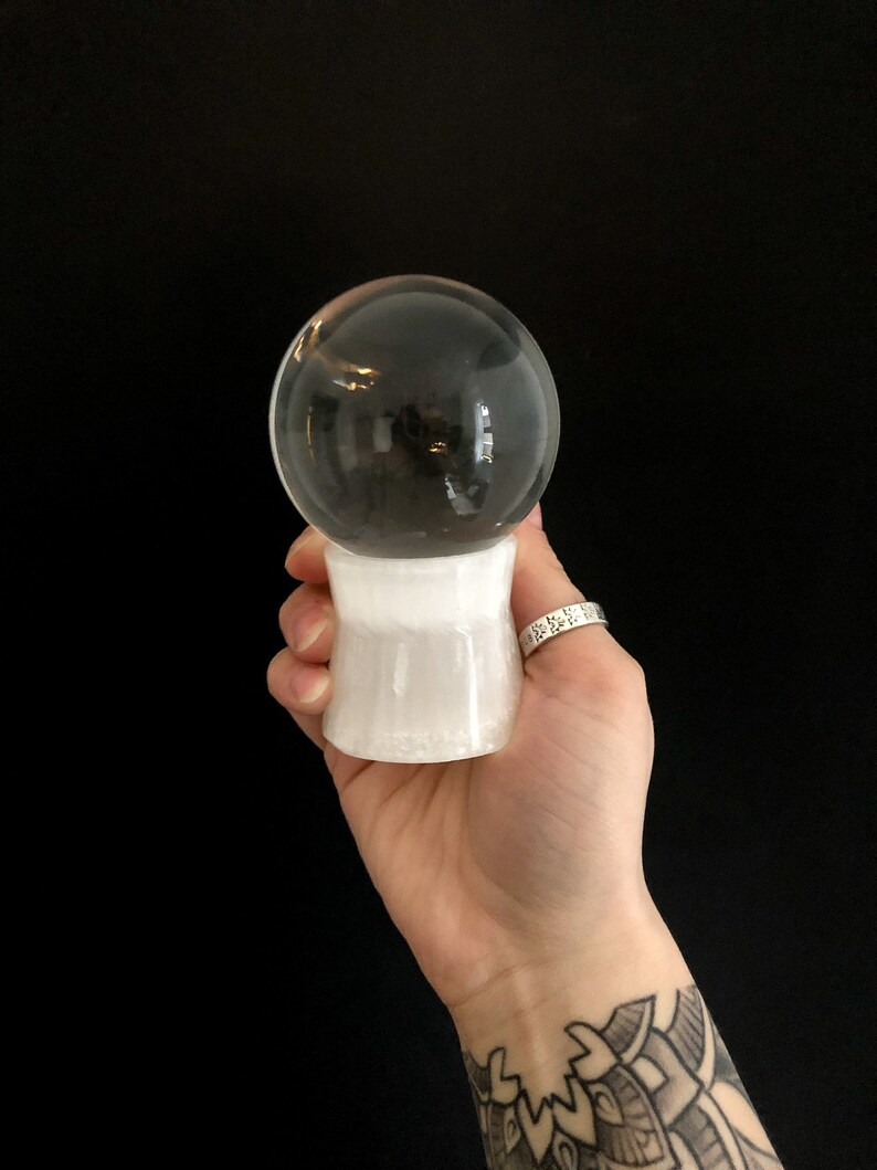 Puede incluir: Una bola de cristal transparente sobre una base cil&iacute;ndrica blanca. La esfera refleja el entorno. La base se sostiene en una mano sobre un fondo negro. La bola de cristal es una herramienta de adivinaci&oacute;n.