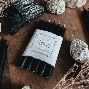 May include: A bundle of black ritual candles wrapped in white paper with the word "Vetiver" printed on it. The Stone Maidens Ritual Candles are displayed on a wooden surface with black tourmaline crystals and other decorative stones.