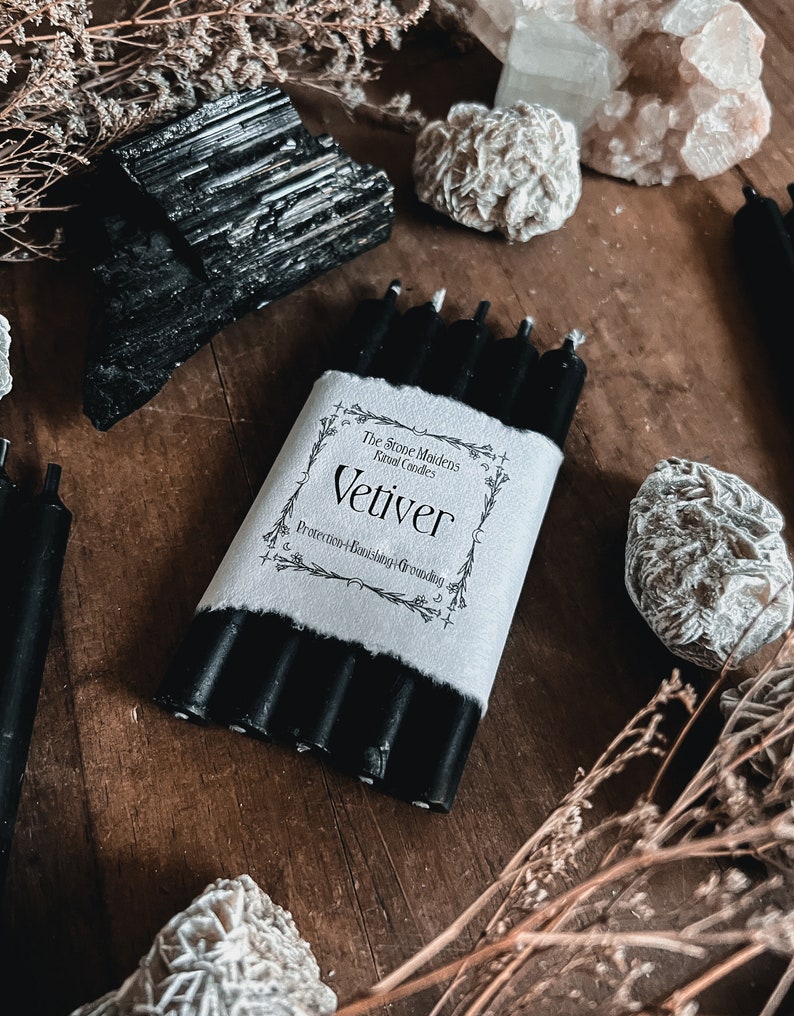 May include: A bundle of black ritual candles wrapped in white paper with the word "Vetiver" printed on it. The Stone Maidens Ritual Candles are displayed on a wooden surface with black tourmaline crystals and other decorative stones.