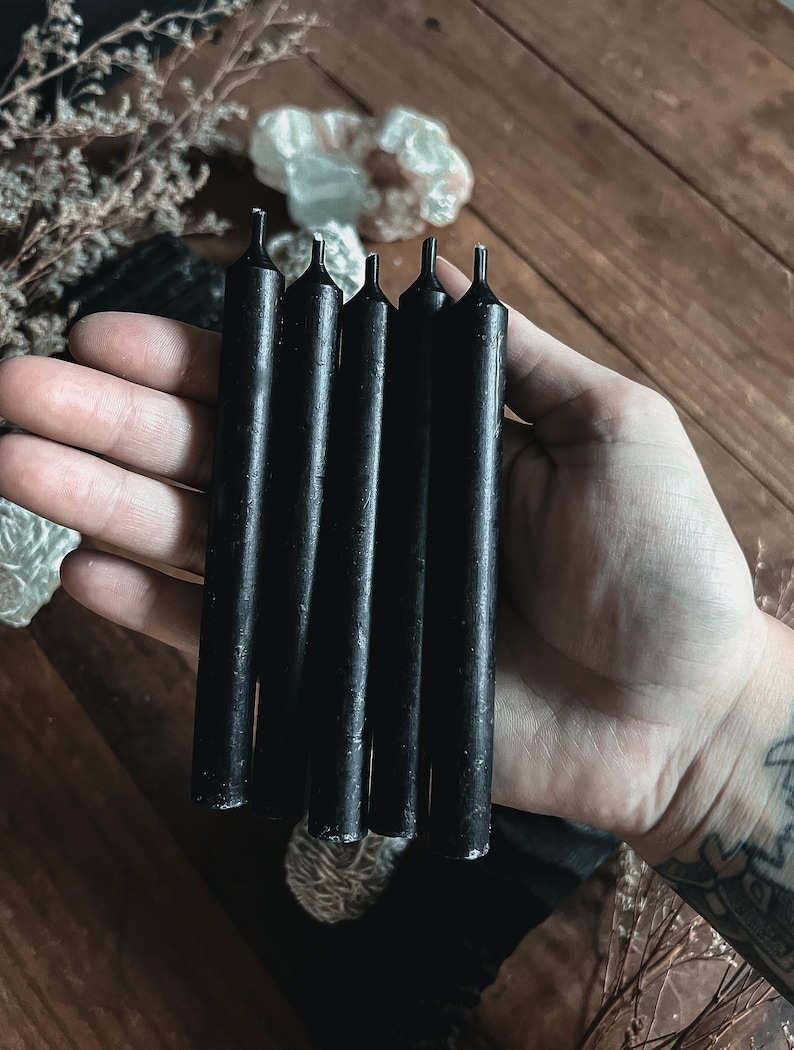 May include: A hand holds five black taper candles. The candles are cylindrical with a slightly textured surface and a small, pointed tip. The background includes a wooden surface and decorative elements.