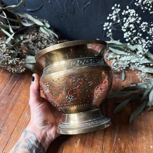 May include: A brass mortar with a wide, flared rim and a pedestal base. The exterior surface features intricate, etched floral patterns and decorative bands, with recessed areas filled with a dark red pigment to highlight the detailed metalwork.