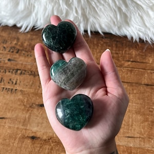Puede incluir: Tres cristales verdes en forma de corazón. Los cristales están pulidos y tienen un color verde oscuro con vetas verdes más claras.