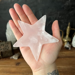 Puede incluir: Un cristal de selenita de color rosa pálido en forma de estrella. El cristal es liso y pulido, con una apariencia ligeramente translúcida.
