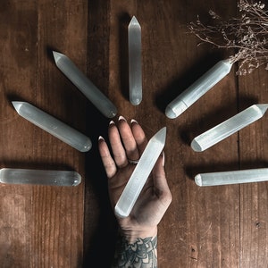 May include: A collection of eight white selenite wands arranged in a circle on a wooden surface. A hand is holding one of the wands in the centre of the circle.