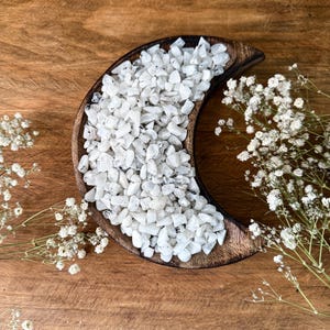 Könnte beinhalten: Eine halbmondförmige Holzschale gefüllt mit kleinen, weißen und grauen Trommelsteinen. Die Schale steht auf einer Holzoberfläche, mit Zweigen weißer Schleierkrautblüten, die darum angeordnet sind. Die Steine scheinen Mondstein zu sein.