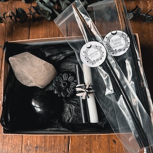 Könnte beinhalten: Geschenkbox mit Räucherstäbchen, Kristallen und Kerzen. Die Räucherstäbchen befinden sich in einer transparenten Verpackung mit Etiketten mit der Aufschrift "Frankincense" und "Dragon's Blood Incense Sticks". Die Box ist mit schwarzem Seidenpapier ausgekleidet.