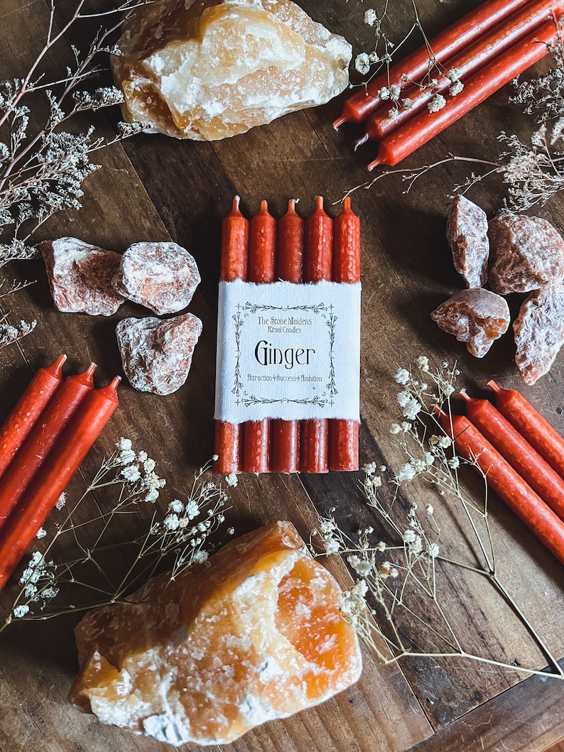 K&ouml;nnte beinhalten: Ein Set mit sechs roten Ingwerkerzen mit einem wei&szlig;en Etikett, auf dem "The Stone Maidens Ritual Candles Ginger" und "For Victory, Success & Abundance" steht. Die Kerzen sind von orangefarbenen Kristallen und wei&szlig;en Blumen umgeben.