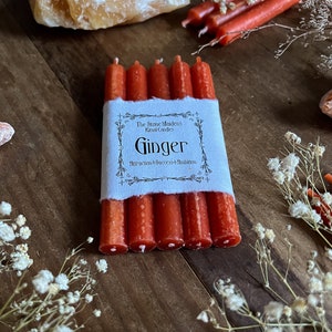 K&ouml;nnte beinhalten: Sechs rote Ingwer-Ritualkerzen, die mit einem wei&szlig;en Etikett zusammengebunden sind, auf dem "The Stone Maidens Ritual Candles Ginger Attraction + Success + Ambition" steht.