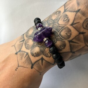 Puede incluir: Un brazalete de roca de lava negra con un gran amuleto de cristal de amatista púrpura. El brazalete se lleva en la muñeca.