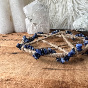May include: A blue and brown crystal grid with a rope base. The grid is made of small blue stones and is surrounded by a thick rope. The grid is sitting on a wooden surface.