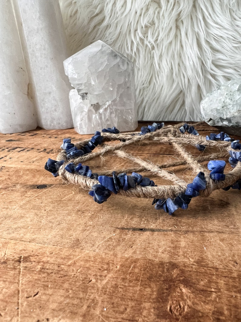 May include: A blue and brown crystal grid with a rope base. The grid is made of small blue stones and is surrounded by a thick rope. The grid is sitting on a wooden surface.