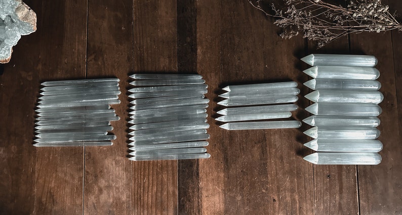May include: A collection of selenite wands, also known as selenite sticks, arranged on a wooden surface. The wands are various lengths and are all white and translucent.