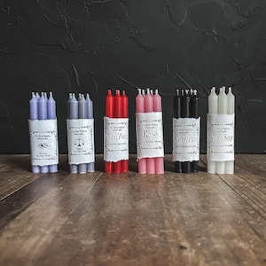 May include: Bundles of ritual candles in various colors, including lavender, gray, red, pink, black, and white. Each bundle is wrapped with a label featuring text. The candles are arranged on a wooden surface against a dark background.