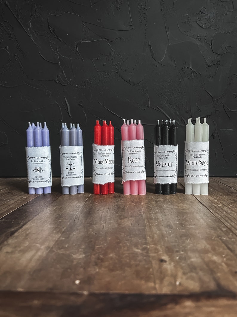 May include: Bundles of ritual candles in various colors, including lavender, gray, red, pink, black, and white. Each bundle is wrapped with a label featuring text. The candles are arranged on a wooden surface against a dark background.