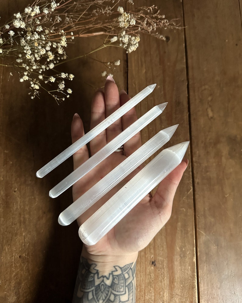 May include: Four white selenite wands of varying lengths, with pointed ends, held in a hand.