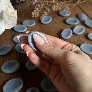 May include: A collection of light blue oval-shaped smooth stones, also known as Celestite palm stones, on a wooden surface. One stone is being held in a hand.
