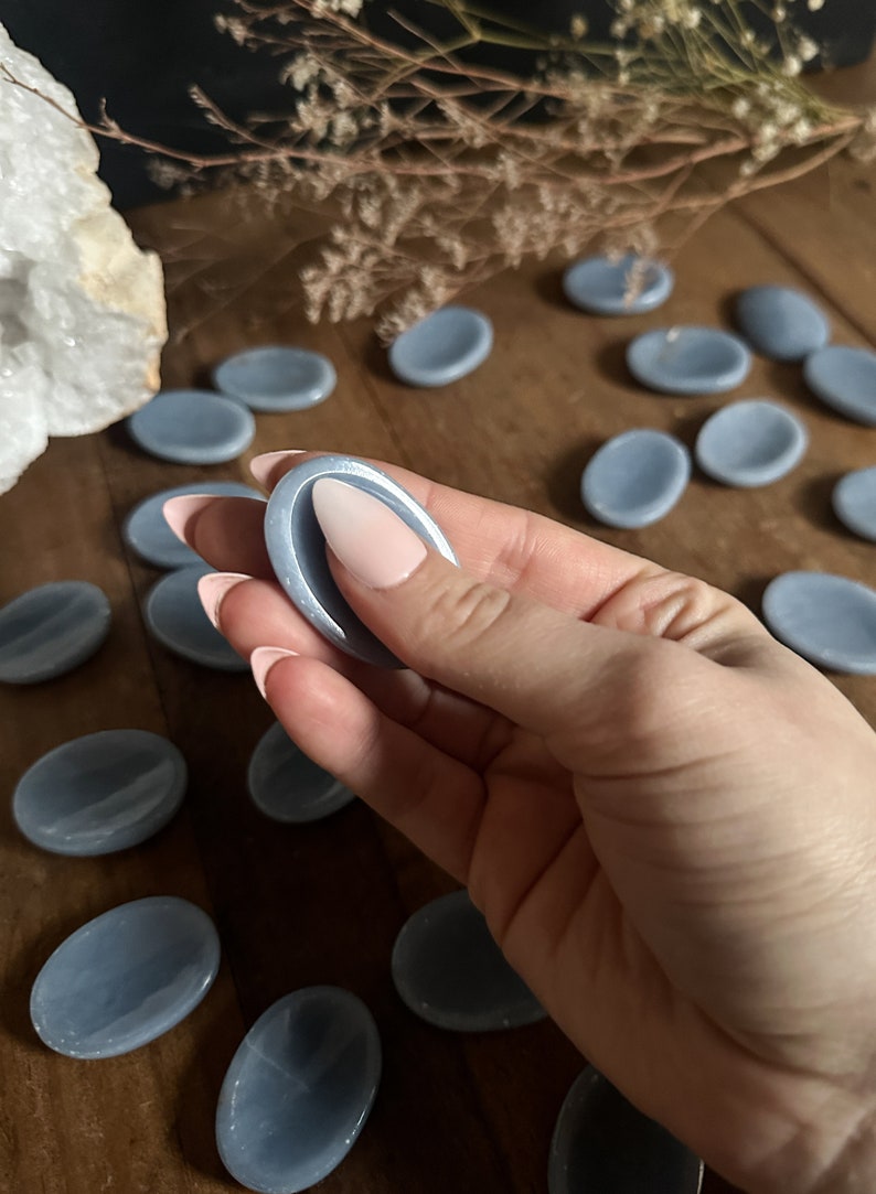 May include: A collection of light blue oval-shaped smooth stones, also known as Celestite palm stones, on a wooden surface. One stone is being held in a hand.
