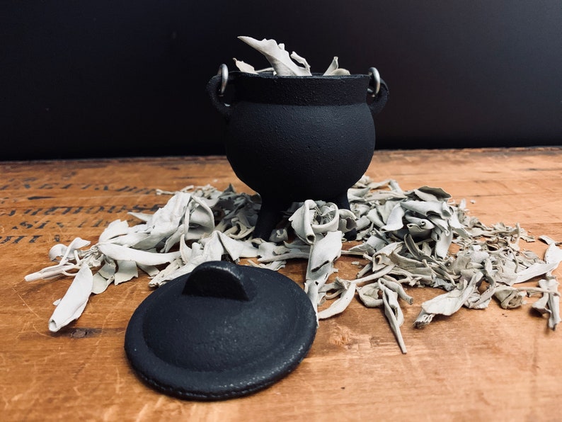 May include: A black cast iron cauldron with a lid, filled with white sage bundles. The cauldron is sitting on a wooden surface with more sage bundles scattered around it.