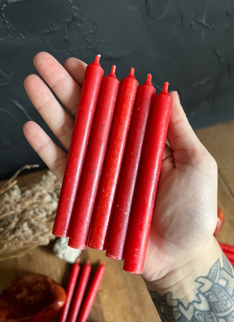May include: A hand holds six red taper candles. The candles are cylindrical, with a slightly textured surface and a wick at the top. The candles are approximately 5 inches long.