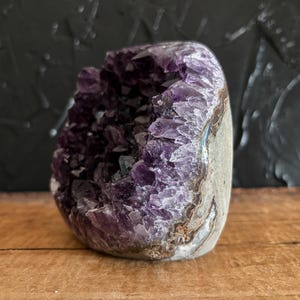May include: A raw amethyst geode featuring deep purple, crystalline structures within a hollow, rounded stone. The exterior displays a natural, textured grey and brown rock surface, while the interior reveals jagged, translucent violet points.