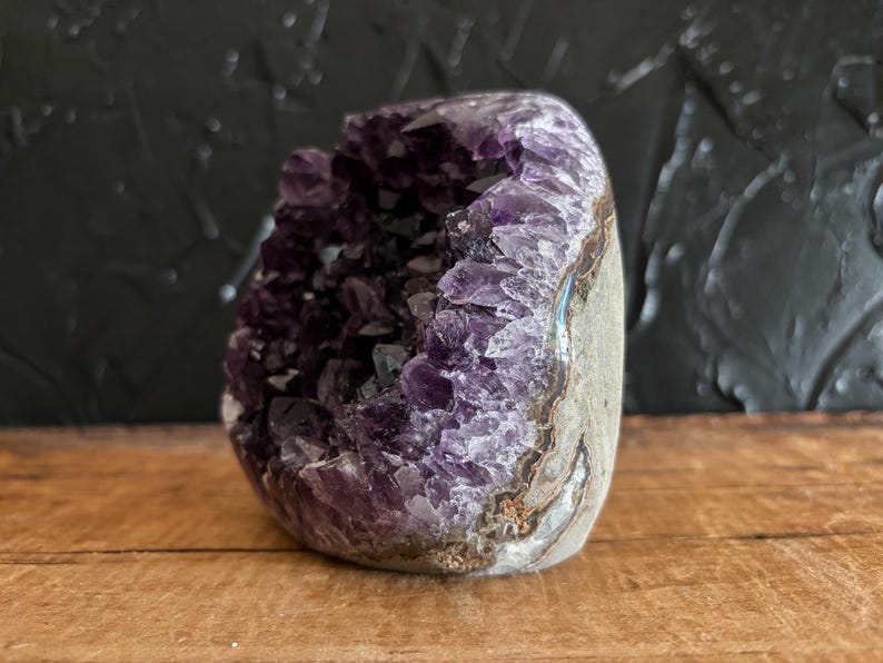 May include: A raw amethyst geode featuring deep purple, crystalline structures within a hollow, rounded stone. The exterior displays a natural, textured grey and brown rock surface, while the interior reveals jagged, translucent violet points.