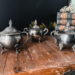 Puede incluir: Tres azucareros de plata con tapas y patas ornamentadas, sentados sobre una superficie de madera.