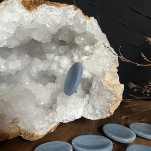 May include: A set of six light blue oval-shaped stones, also known as gua sha stones, are arranged on a wooden surface. One stone is resting inside a large white crystal geode.