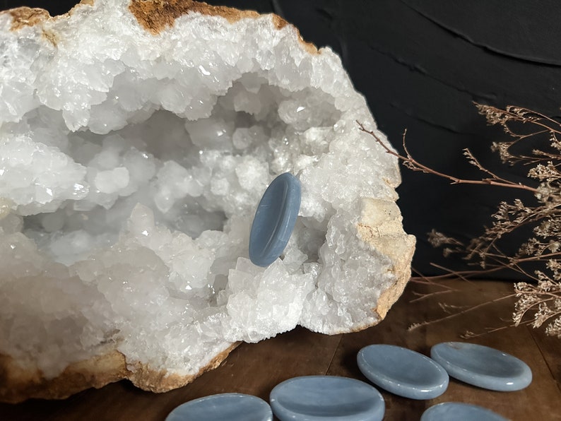 May include: A set of six light blue oval-shaped stones, also known as gua sha stones, are arranged on a wooden surface. One stone is resting inside a large white crystal geode.