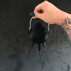 May include: A small black cauldron with a silver handle, held by a hand against a dark gray textured background.
