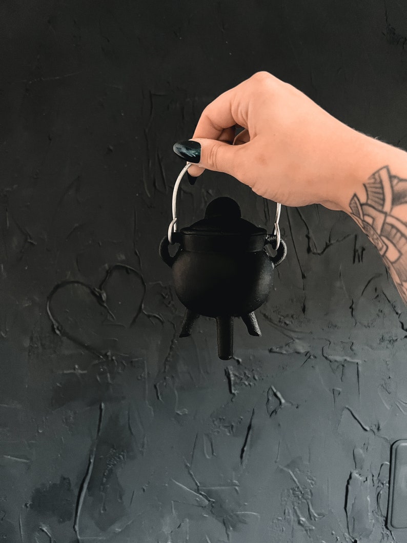 May include: A small black cauldron with a silver handle, held by a hand against a dark gray textured background.