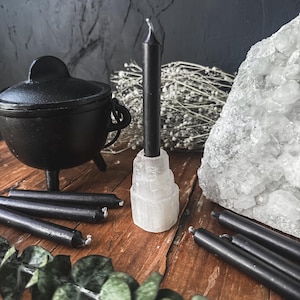 May include: A collection of black taper candles, a small black cauldron, and a large white crystal cluster arranged on a wooden surface. The candles are of varying lengths, some standing upright in a selenite holder. Dried flowers and greenery add to the display.