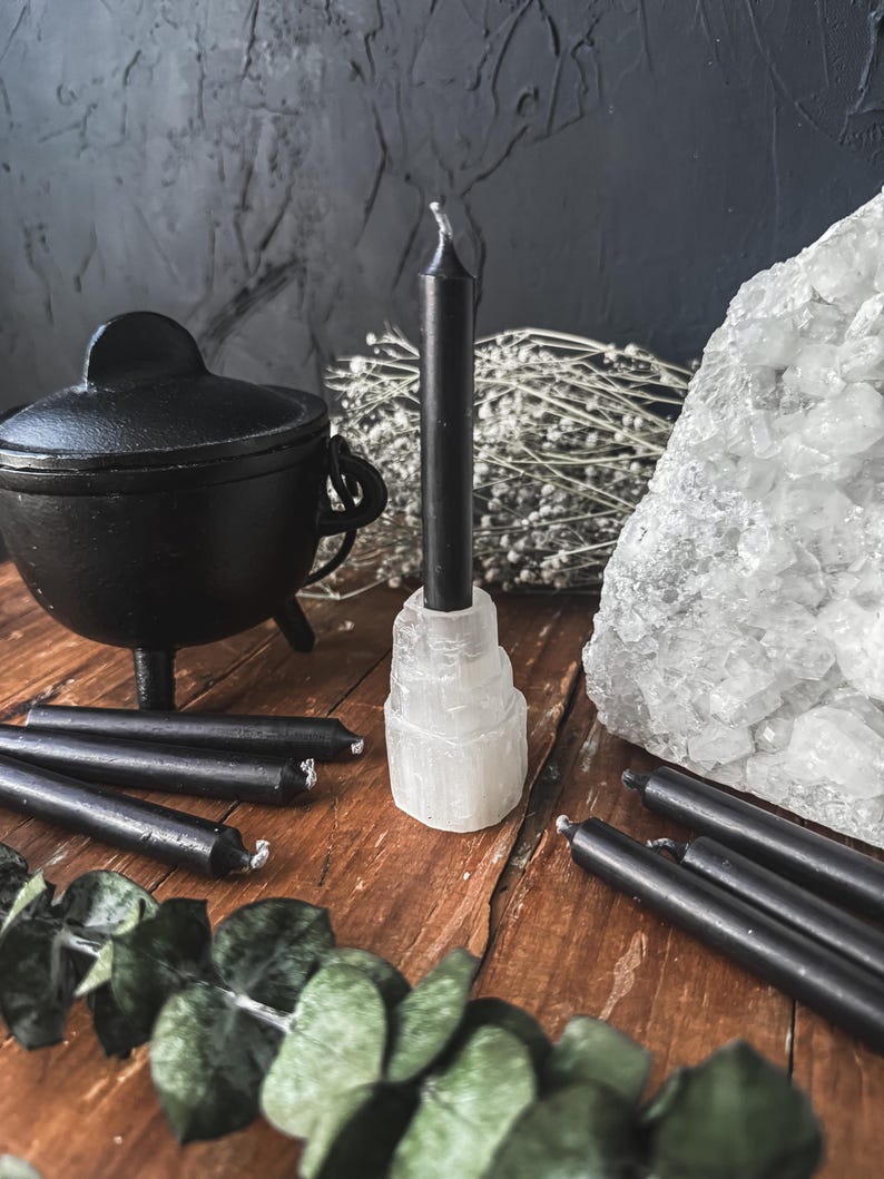 May include: A collection of black taper candles, a small black cauldron, and a large white crystal cluster arranged on a wooden surface. The candles are of varying lengths, some standing upright in a selenite holder. Dried flowers and greenery add to the display.