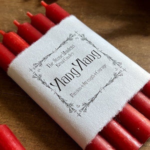 May include: A bundle of red ritual candles wrapped in white paper. The paper is printed with the words "Ylang Ylang" and "Passion + Strength + Courage". The brand name "The Stone Maidens Ritual Candles" is also printed on the paper.
