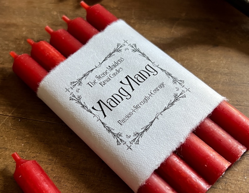May include: A bundle of red ritual candles wrapped in white paper. The paper is printed with the words "Ylang Ylang" and "Passion + Strength + Courage". The brand name "The Stone Maidens Ritual Candles" is also printed on the paper.