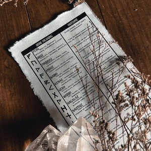 May include: A black and white chart with the names of runes and their meanings and outcomes. The chart is on a wooden surface with dried flowers.