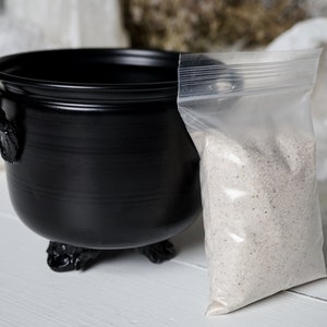 May include: A black cauldron with three legs and a handle sits on a white surface next to a clear plastic bag filled with white sand.