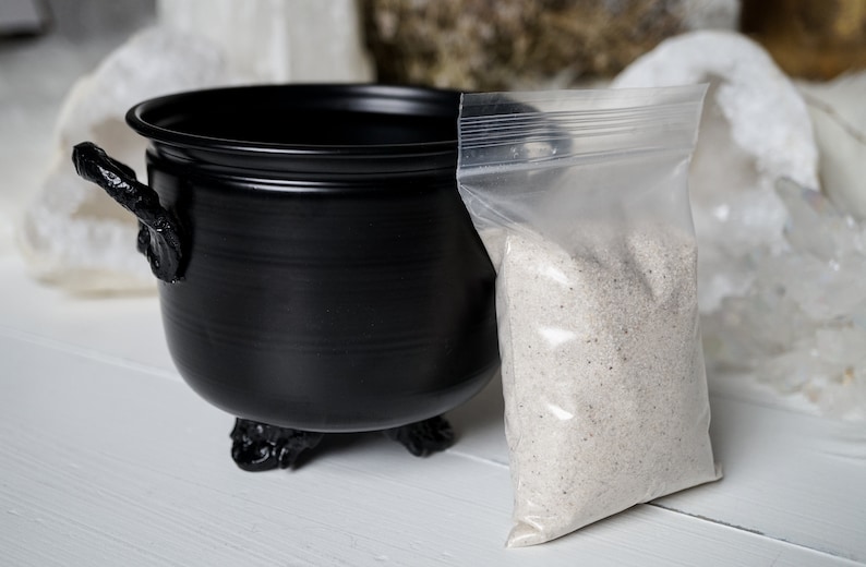 May include: A black cauldron with three legs and a handle sits on a white surface next to a clear plastic bag filled with white sand.