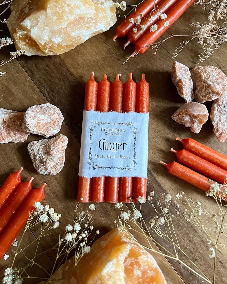 K&ouml;nnte beinhalten: Ein Set mit sechs orangefarbenen Ingwer-Ritualkerzen mit einem wei&szlig;en Etikett, auf dem "The Stone Minders Ritual Candles Ginger  Attraction/Success/Abundance" steht.