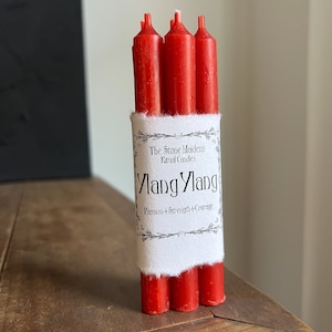 May include: A bundle of three red ritual candles. The candles are wrapped with a white label that reads "Ylang Ylang, Passion + Strength + Courage" and "The Stone Maidens Ritual Candles". The candles are on a wooden surface.